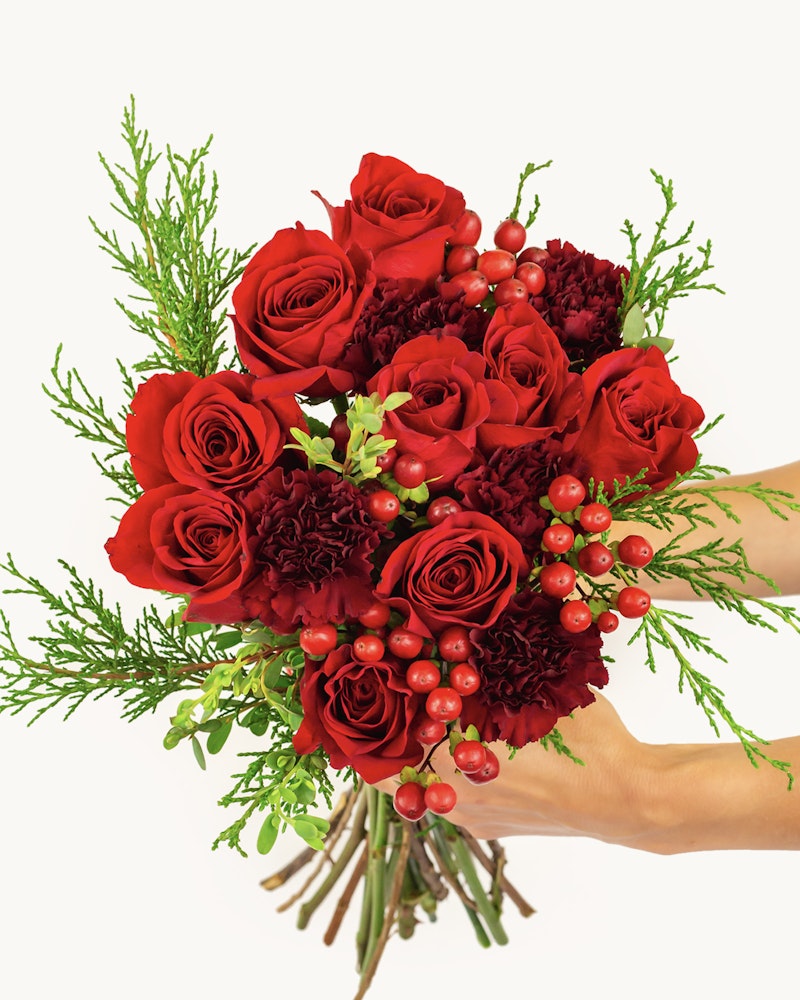 A close-up view of a person's hand holding a beautiful bouquet of dark red roses, carnations and hypericum berries with greenery accents on a white background.