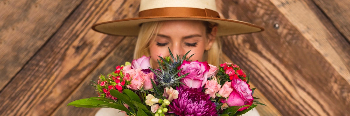 A woman holds a vibrant floral bouquet featuring pink and purple blooms, wearing a stylish hat.