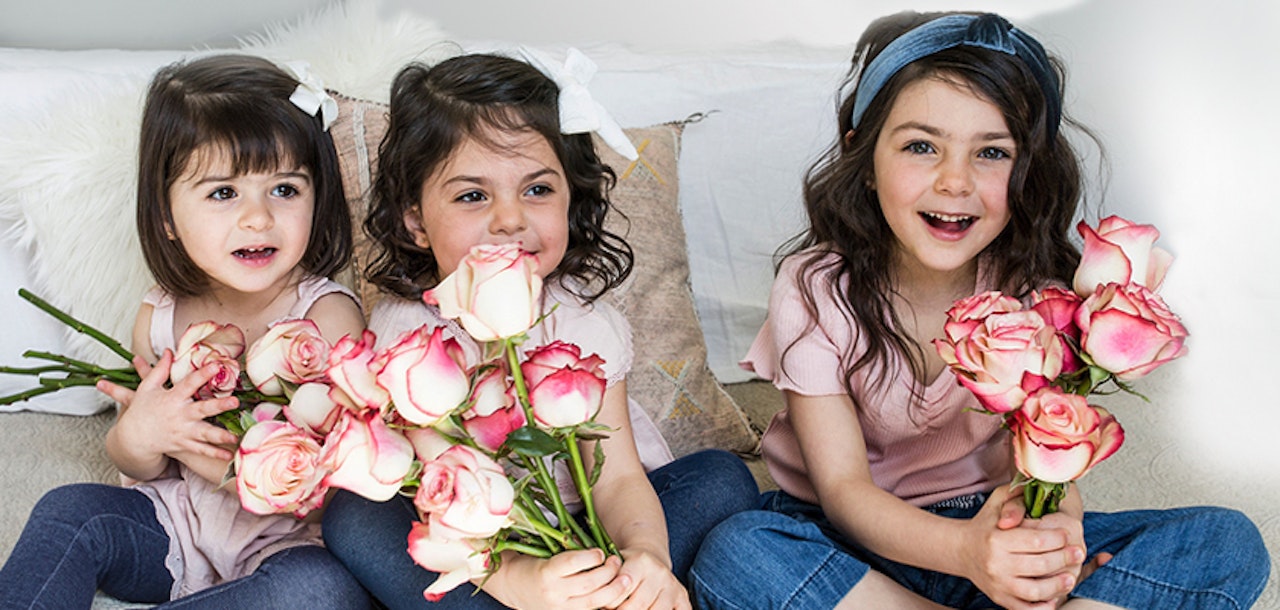 Three smiling young sisters sitting together on a sofa, each holding a bouquet of pink roses, wearing casual outfits with adorable white bows in their hair.