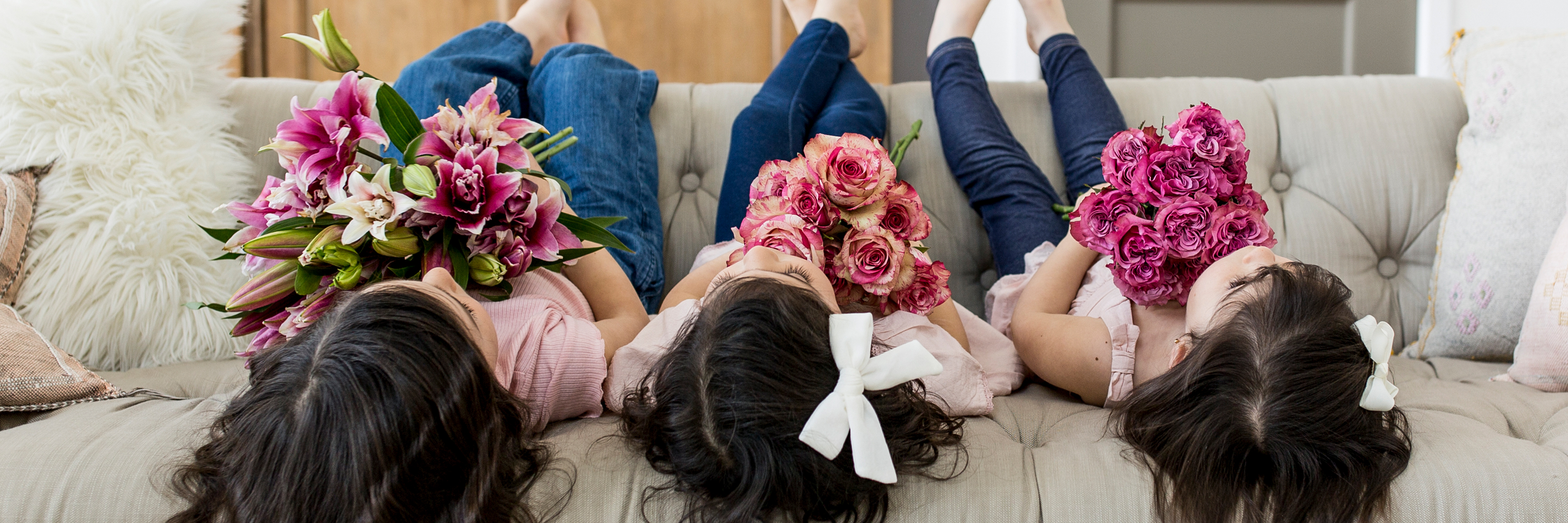Three girls lounging on a couch, each holding vibrant floral bouquets, smiling joyfully.