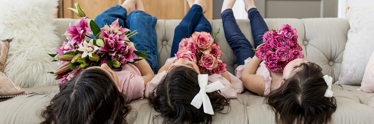 Three girls lounging on a couch, each holding vibrant floral bouquets, smiling joyfully.