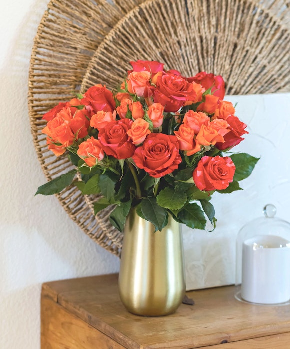 A bouquet of vibrant orange roses arranged in a gold vase on a wooden table, with a woven circular wall decor in the background, creating a warm, homey atmosphere.
