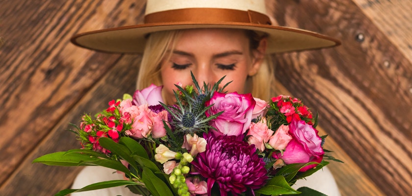 A woman in a wide-brimmed hat holds a vibrant floral bouquet, enjoying its fragrance.
