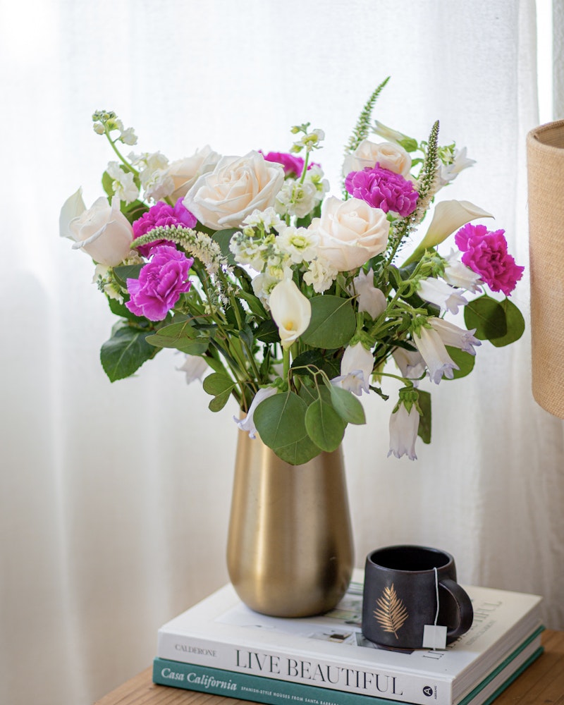 Elegant bouquet of white roses, purple flowers, and greenery in a gold vase on a stack of lifestyle books beside a black mug on a wooden table with a sheer curtain background.