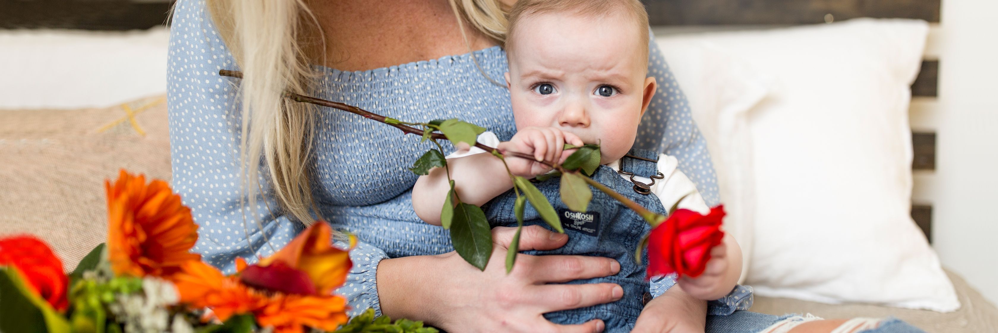 A cute baby holding a rose while sitting beside a vibrant flower arrangement.