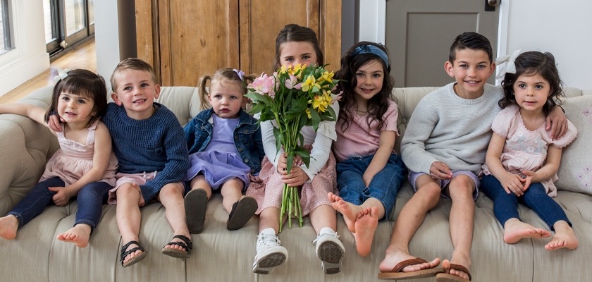 A cheerful group of seven children sitting on a sofa, smiling and enjoying a moment together.