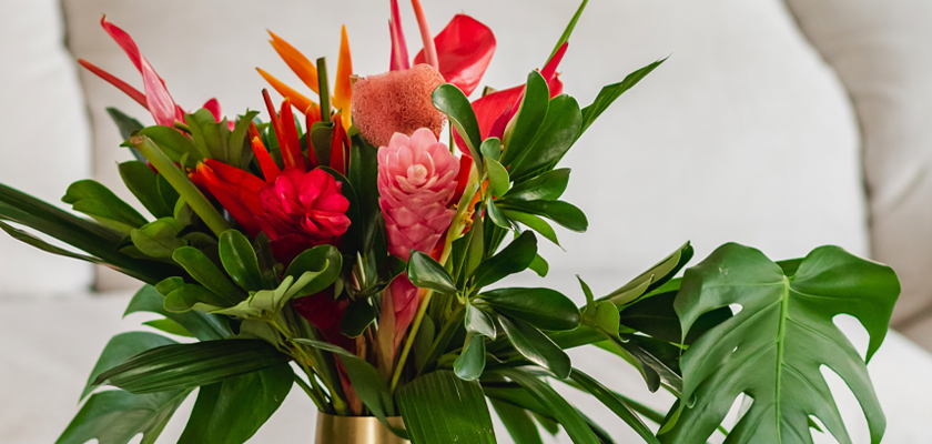 Vibrant tropical bouquet featuring red and pink flowers and lush green foliage in a gold vase.