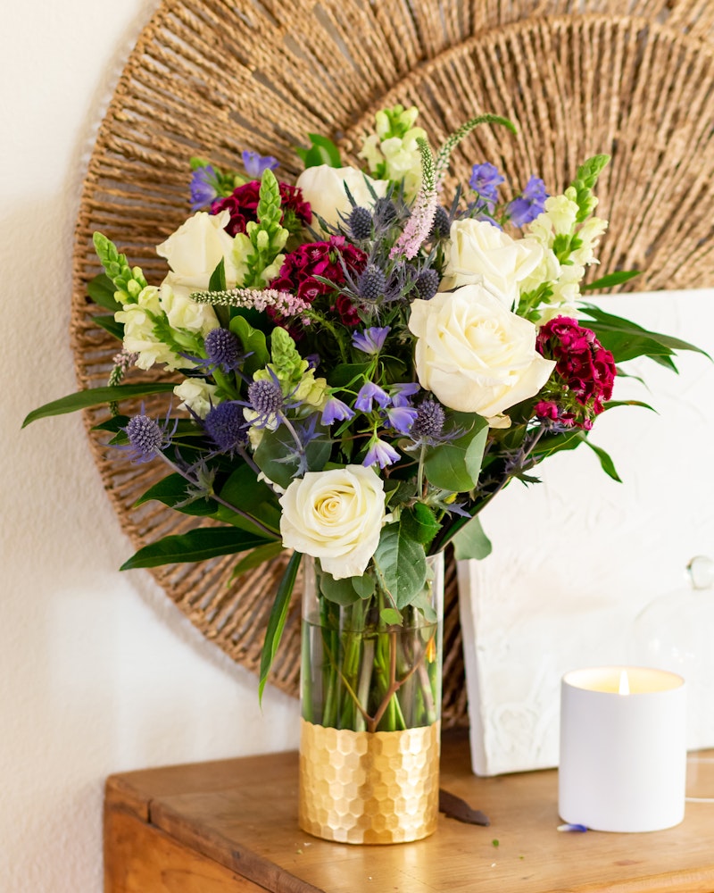 A vibrant bouquet of white roses, purple flowers, and greenery arranged in a golden vase on a wooden surface, with a lit candle and wicker decor in the background.