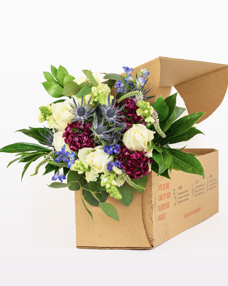 A vibrant bouquet of flowers with purple, white, and green blooms, artistically arranged and presented in an open cardboard box on a white background.
