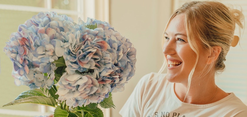 A cheerful woman holding a large bouquet of blue hydrangeas, smiling brightly indoors.