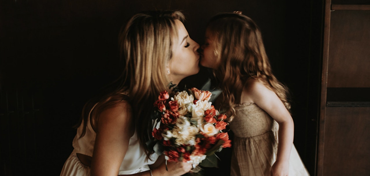 A tender moment captured as a woman leans over to gently kiss a young girl on the lips, both with closed eyes, the woman holding a bouquet of colorful flowers.
