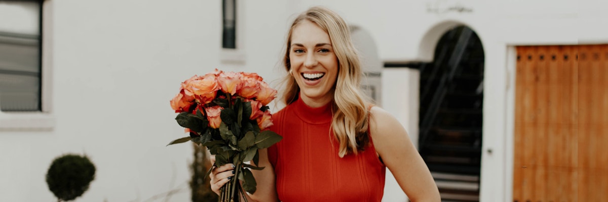 Smiling woman in a red dress joyfully holds a bouquet of vibrant roses outside a home.