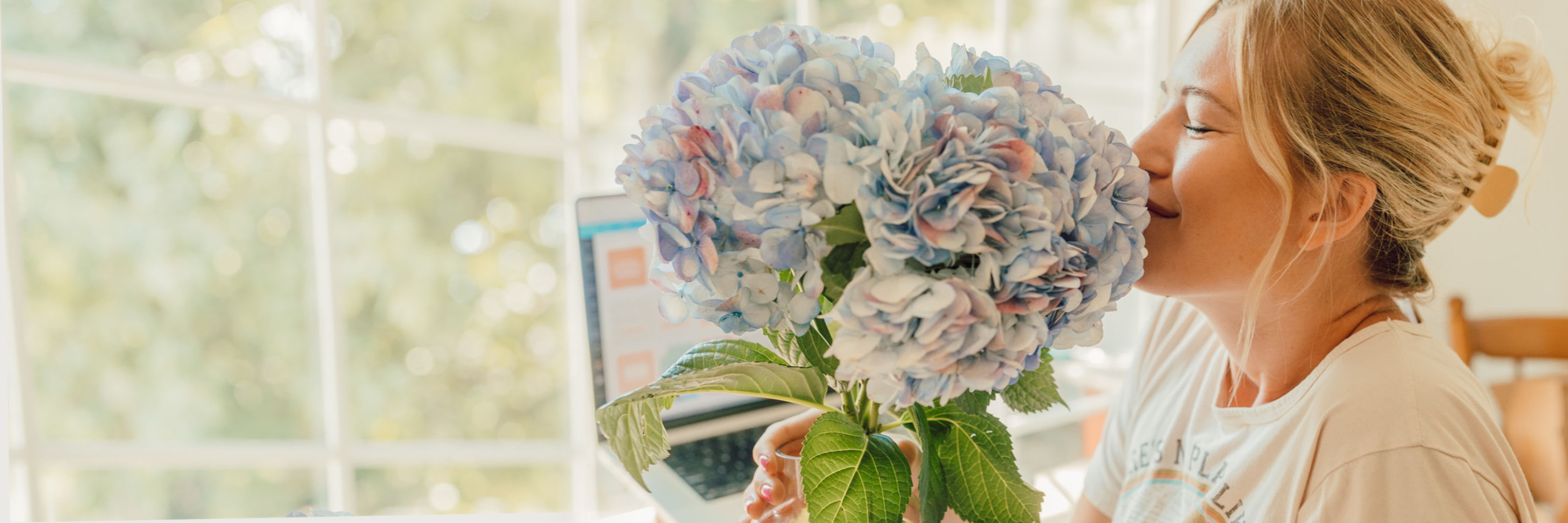 Cheerful woman enjoying a bouquet of blue hydrangeas at her desk, creating a bright atmosphere.