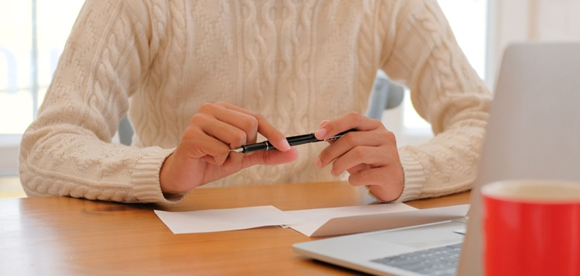 A person in a cozy sweater holding a pen, seated at a desk with a laptop and paper.