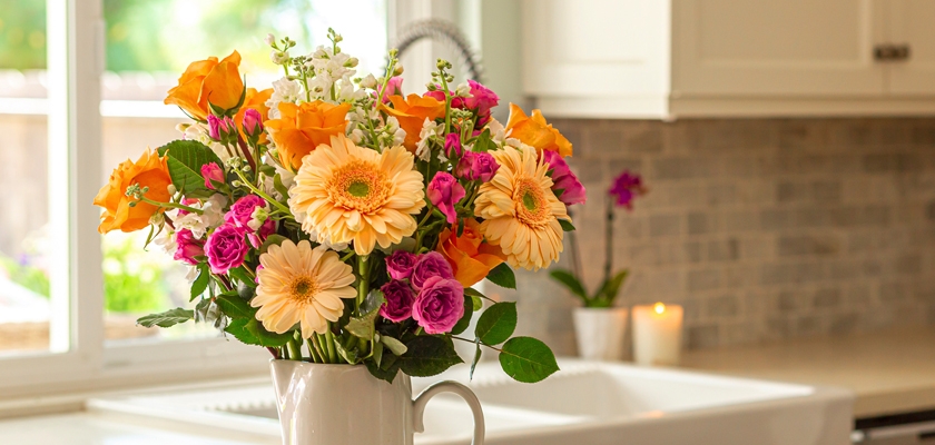 Bright floral arrangement featuring orange roses and gerbera daisies in a white pitcher.