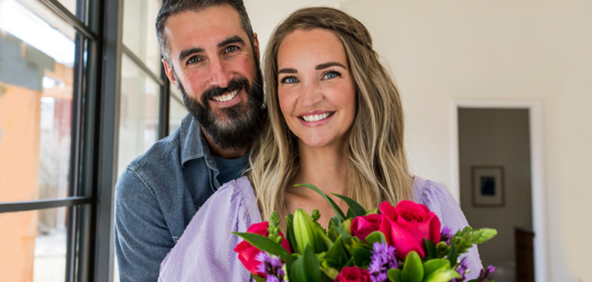 A happy couple poses indoors with a vibrant bouquet of flowers, celebrating love and joy.