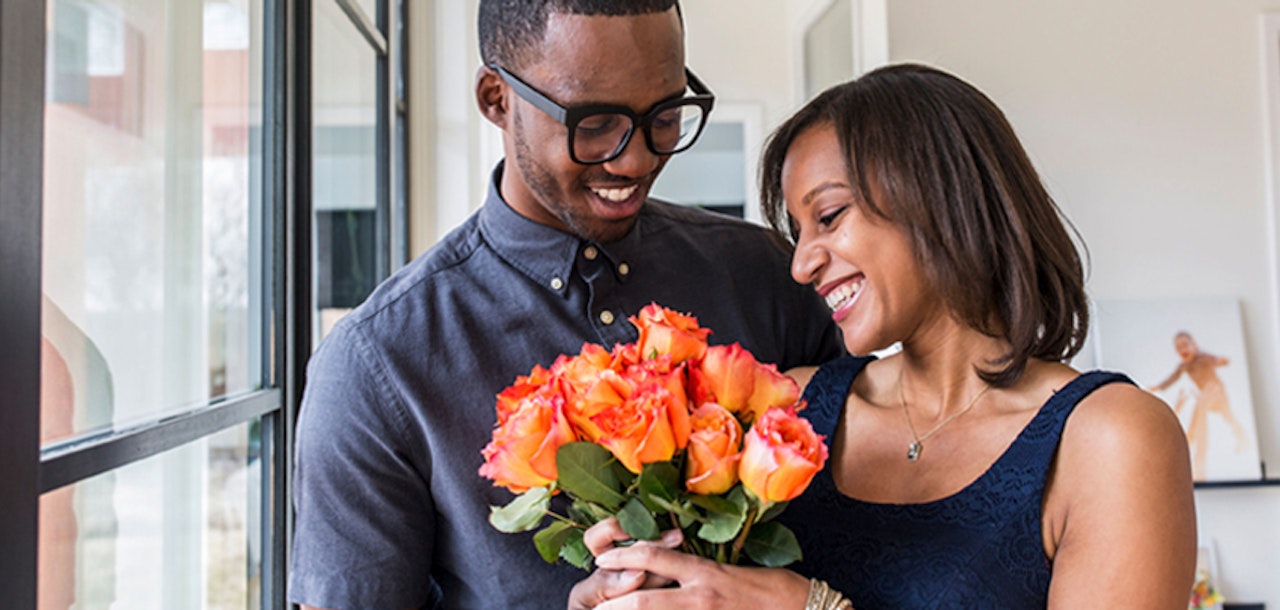A couple joyfully exchanging a vibrant bouquet of orange roses indoors, celebrating love.