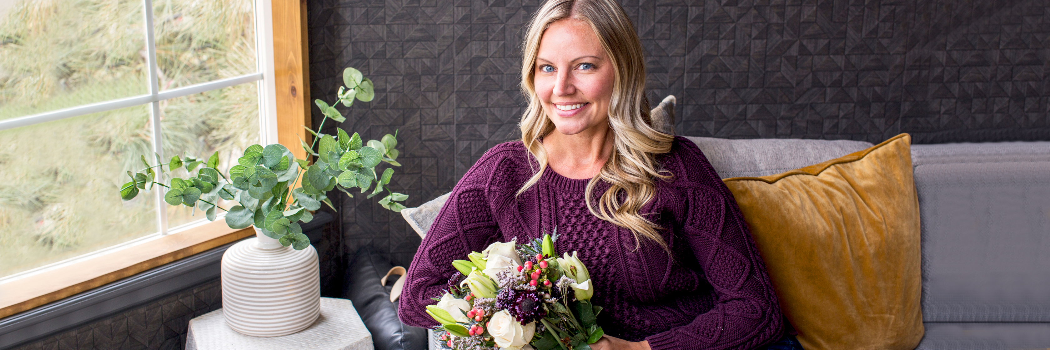 A smiling woman holds a lush bouquet of flowers while sitting by a window, cozy and content.