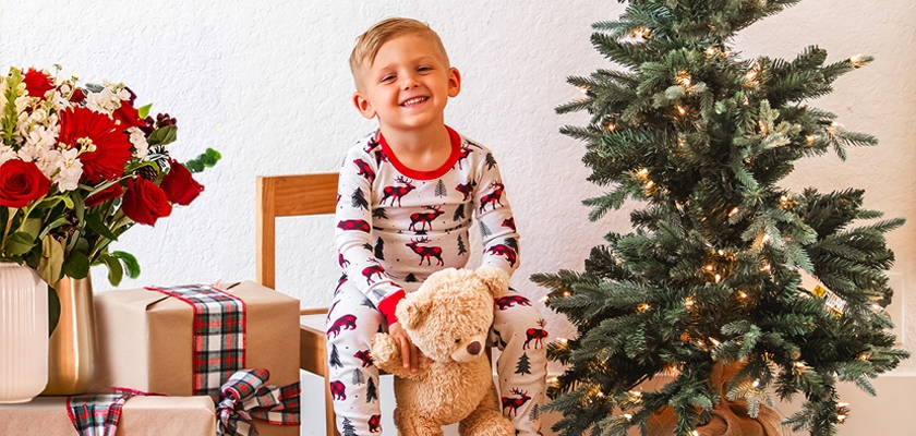 Smiling child in festive pajamas holding a teddy bear beside a Christmas tree and gifts.