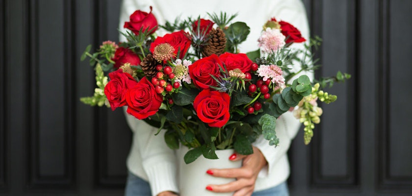 A festive bouquet of red roses, greens, and pinecones in a charming white pot.