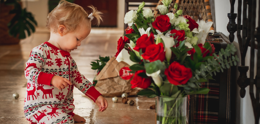 A playful toddler in festive pajamas explores a vibrant bouquet of red roses.