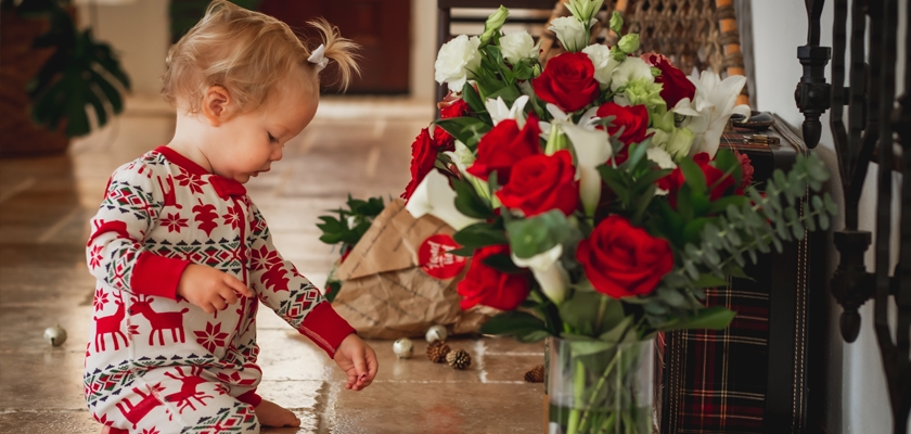 A playful toddler in festive pajamas explores a vibrant bouquet of red roses.