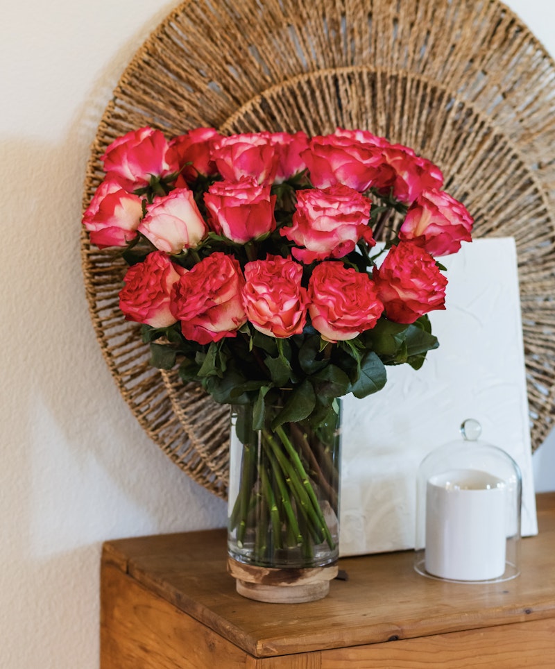 Vibrant bouquet of pink and red roses arranged in a clear glass vase on a wooden table, with a woven wall decoration and a white candle in the background.