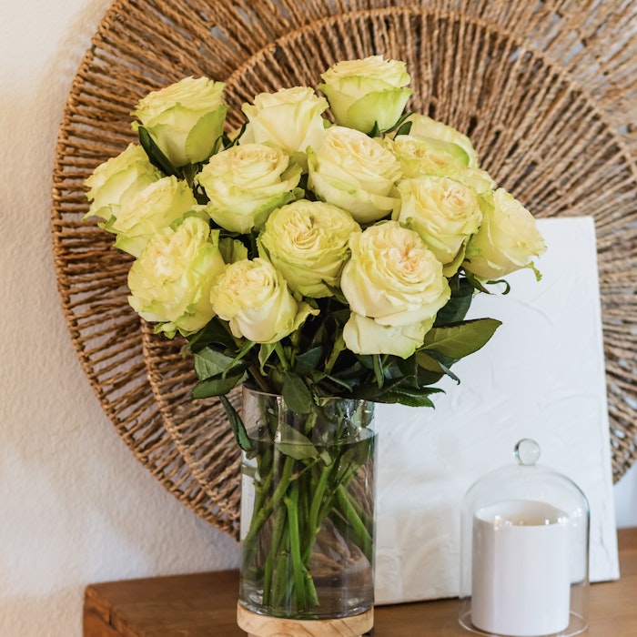 A beautiful bouquet of fresh pale yellow roses in a clear glass vase, set on a wooden sideboard against a textured wall with a woven circular decor piece.