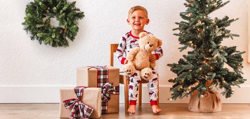 Cheerful child in festive pajamas holding a teddy bear, surrounded by holiday gifts and decorations.