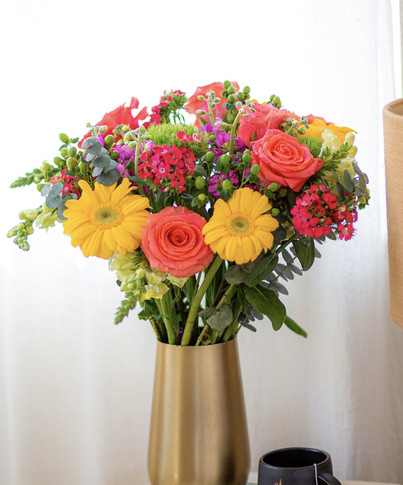 Vibrant bouquet of fresh flowers with yellow gerberas, orange roses, and lush greenery arranged in a sleek gold vase on a light background, symbolizing spring freshness.