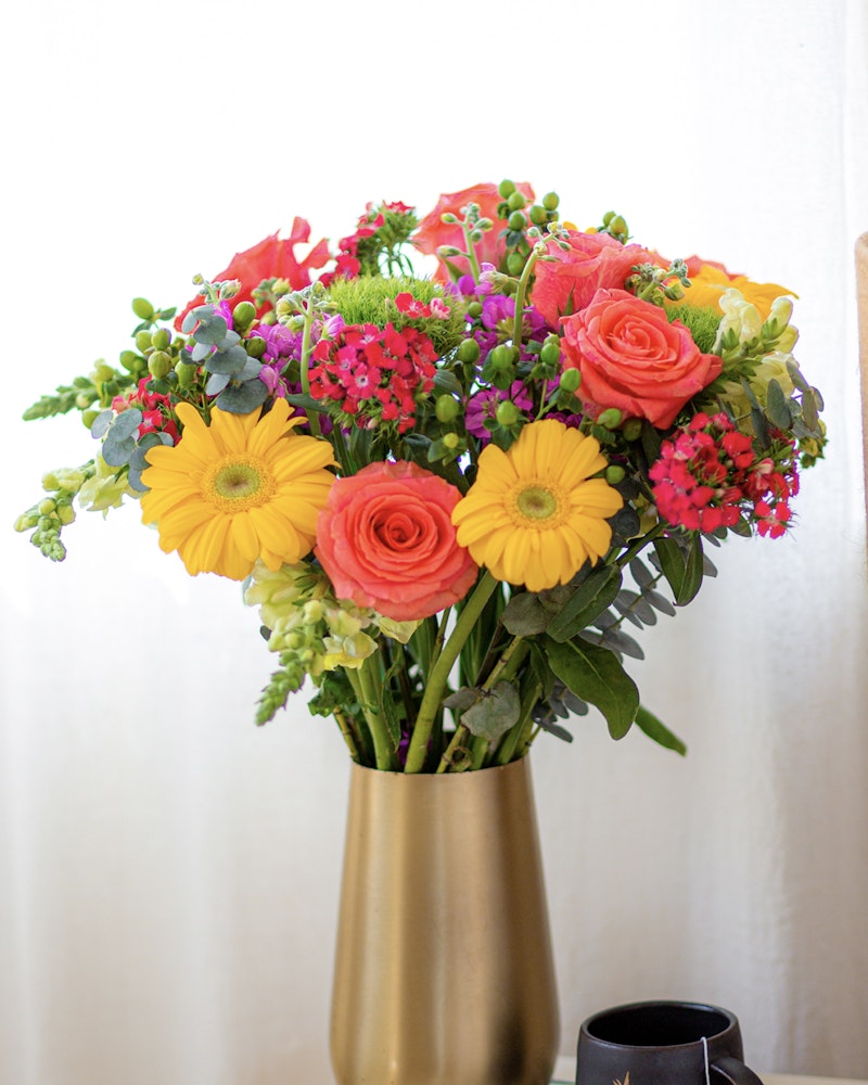 Vibrant bouquet of fresh flowers with yellow gerberas, orange roses, and lush greenery arranged in a sleek gold vase on a light background, symbolizing spring freshness.