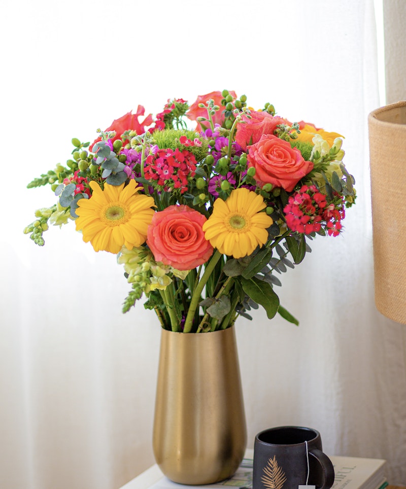 Bright bouquet of fresh flowers with orange roses, yellow gerberas, and purple accents in a gold vase on a table beside a coffee cup with a fern design.