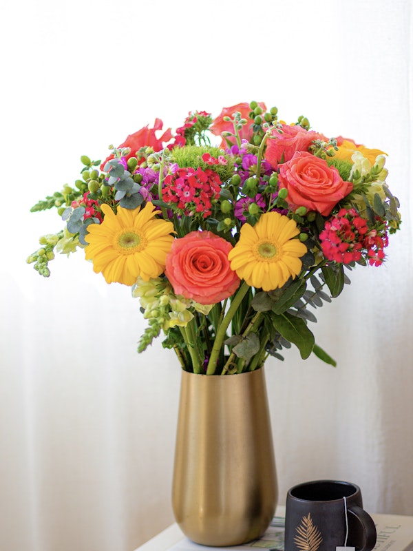 Bright bouquet of fresh flowers with orange roses, yellow gerberas, and purple accents in a gold vase on a table beside a coffee cup with a fern design.