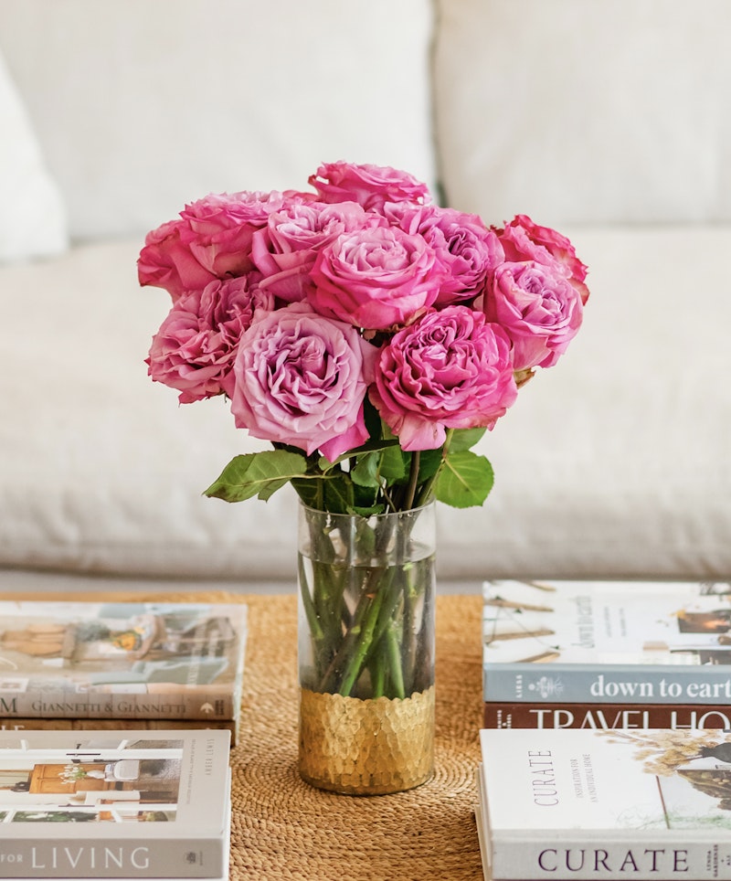Vibrant pink roses arranged in a clear vase on a coffee table, with assorted lifestyle books scattered around, creating an elegant and cozy living room ambiance.