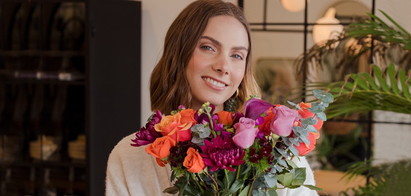 Smiling woman holding a vibrant bouquet of roses and colorful flowers in a cozy setting.