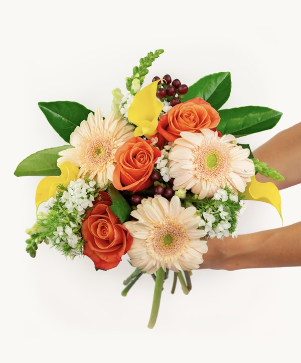A vibrant bouquet of flowers featuring peach gerberas, orange roses, green foliage, and white accent flowers, presented against a white background.