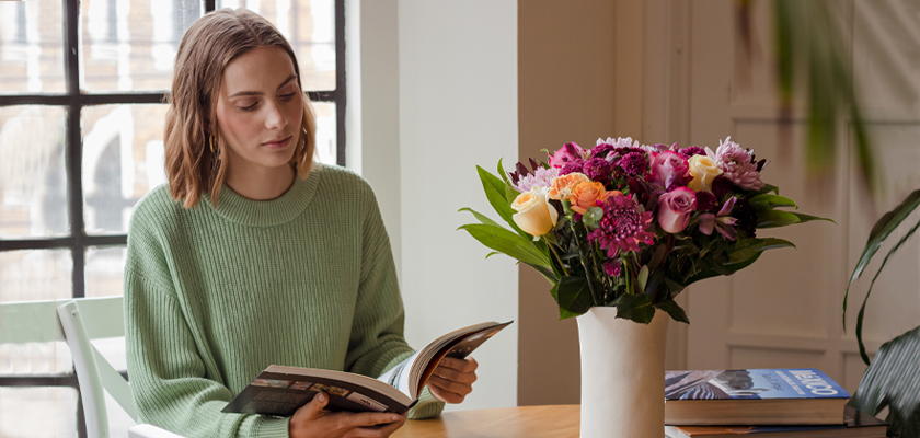 A woman in a cozy sweater reads a book beside a vibrant floral arrangement on a wooden table.