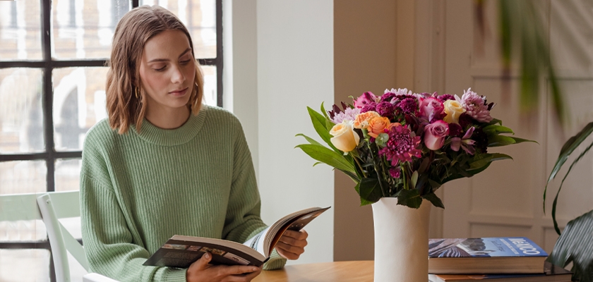 A woman in a cozy sweater reads a book beside a vibrant floral arrangement on a wooden table.