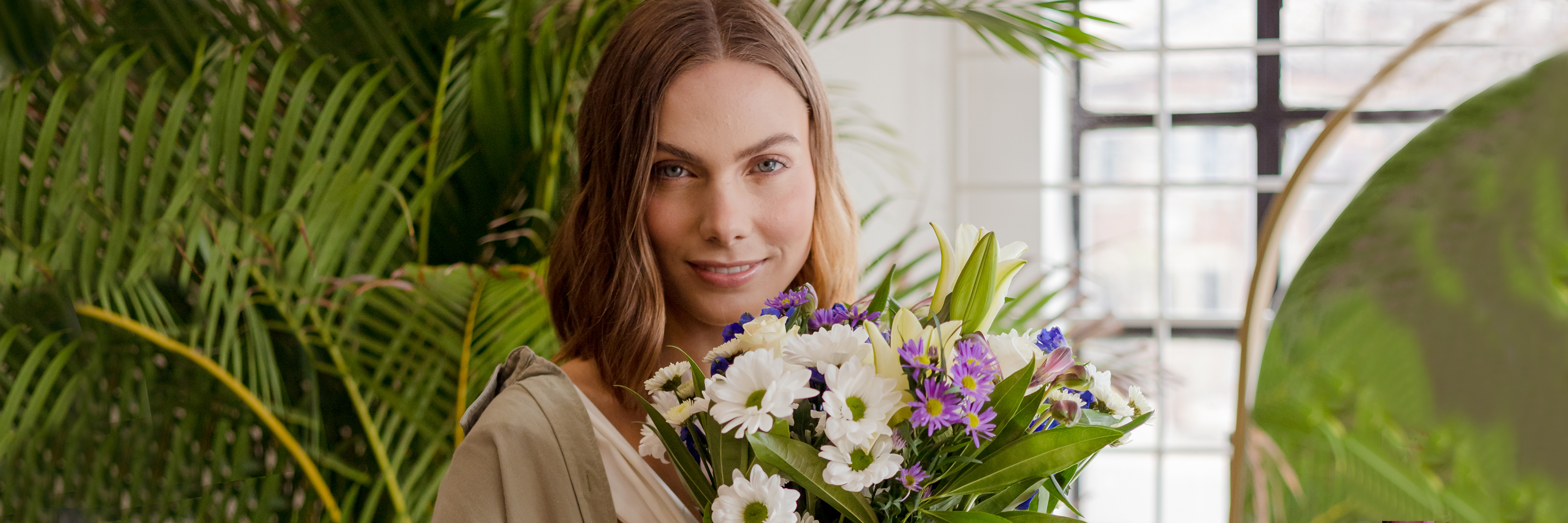 A smiling woman holds a vibrant bouquet of mixed flowers, surrounded by lush greenery.