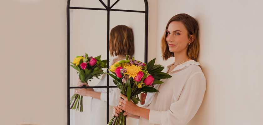 A woman in a white blouse holding a vibrant bouquet of mixed flowers, beside a mirror.