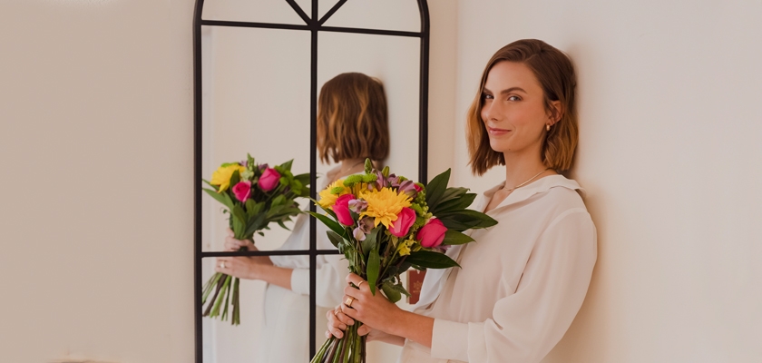 A woman in a white blouse holding a vibrant bouquet of mixed flowers, beside a mirror.