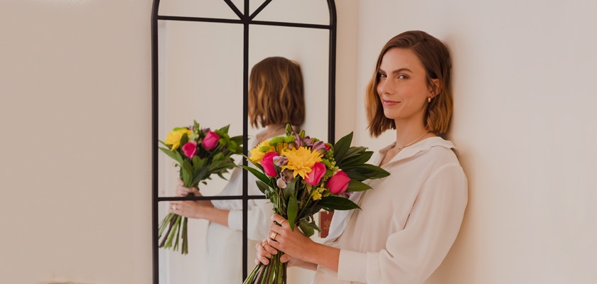 A woman in a white blouse holding a vibrant bouquet of mixed flowers, beside a mirror.