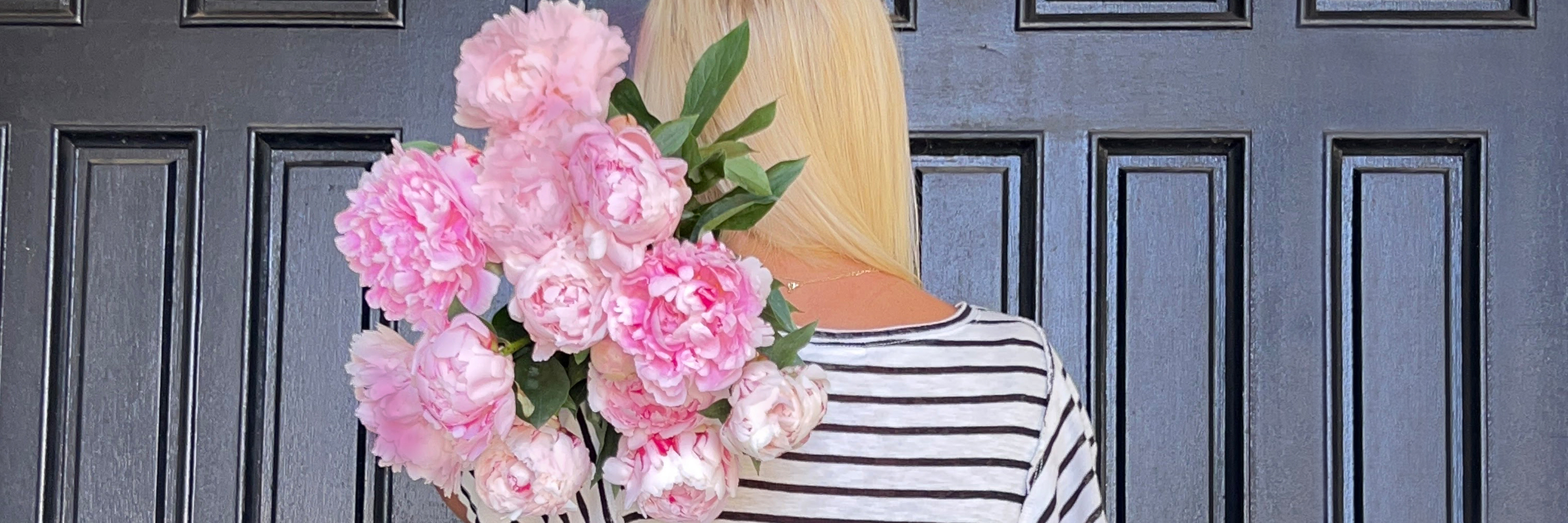 A woman holding a lush pink peony bouquet against a dark wooden door.