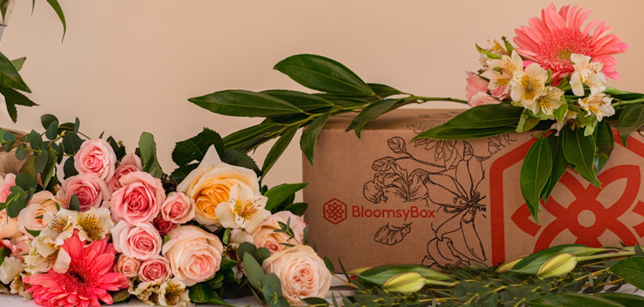 A vibrant arrangement of fresh flowers including pink roses and gerberas beside a BloomsyBox, with a floral illustration on the cardboard packaging.
