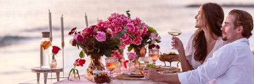 Couple enjoying a romantic beach dinner at sunset, with a table full of flowers, candles, and a fine meal, while holding glasses of wine.