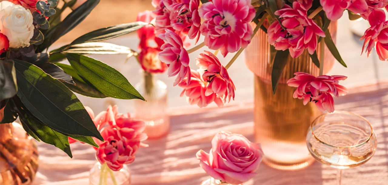 Vibrant pink flowers in a gold vase on a sunlit table with elegant table settings, including a clear glass and delicate floral decorations.