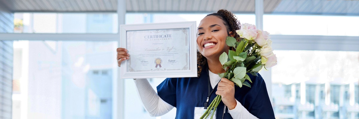 Smiling woman in scrubs holding a diploma and a bouquet of flowers, celebrating achievement.