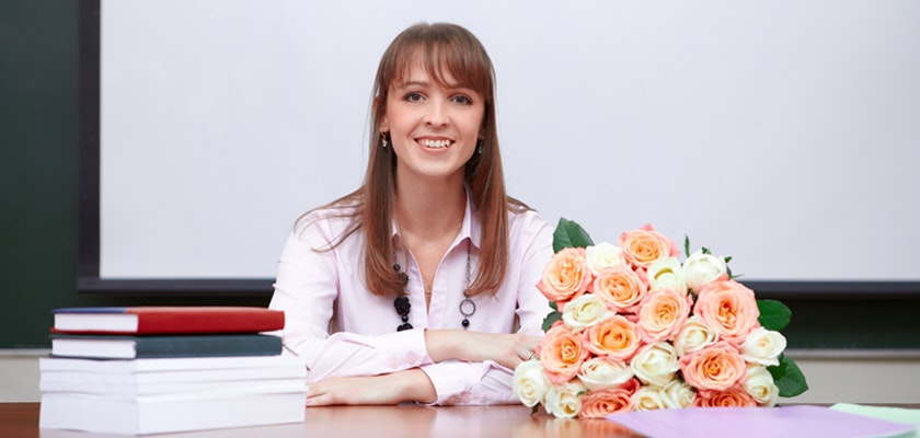 A smiling woman in a classroom setting with a colorful bouquet of roses on her desk.