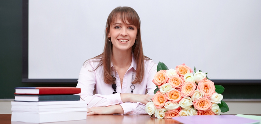 A smiling woman in a classroom setting with a colorful bouquet of roses on her desk.