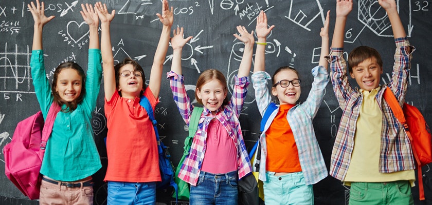 Five cheerful school children raising their hands in excitement, in front of a chalkboard.